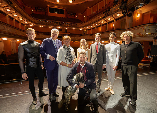 Acht Personen, darunter Frau Herta Stockbauer und Herr Nikolaus Juhász, und ein Hund (in der Mitte) stehen auf der Bühne in Stadttheater. Hinter ihnen die Sitzplätze.