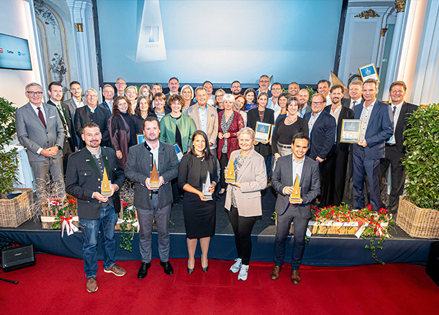 Vielen Menschen stehen auf einem Podium und zeigen ihre Auszeichnung in die Kamera und schauen nach oben.