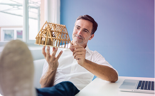 Mann sitzt am Schreibtisch mit einem Modellhaus in der Hand