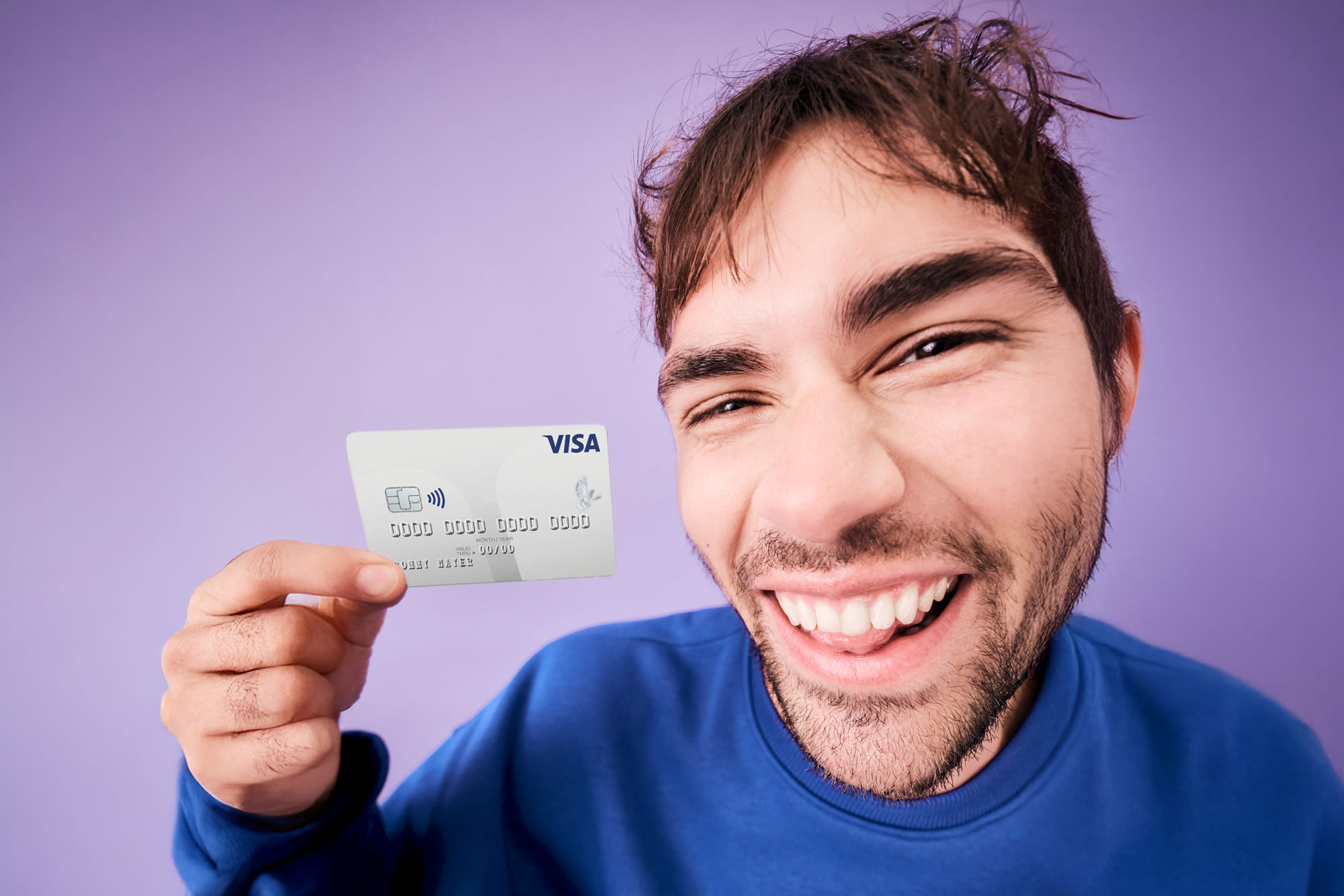 Junger Mann im blauen Pullover zeigt lächelnd seine Visa-Karte vor lila Hintergrund.