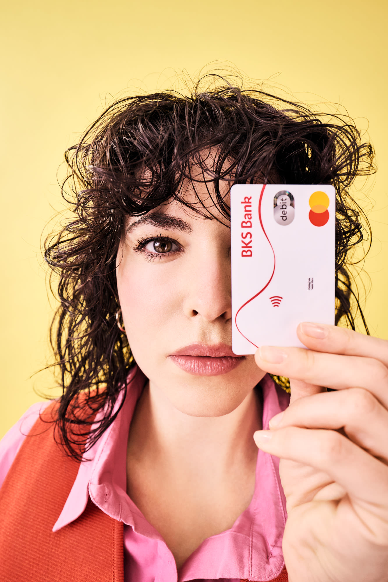 Frau hält BKS Debitkarte direkt vor ihr Gesicht und steht vor gelbem Hintergrund.